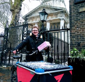 A smiling man stands next to a large black and pink cargo bike with a box on the front